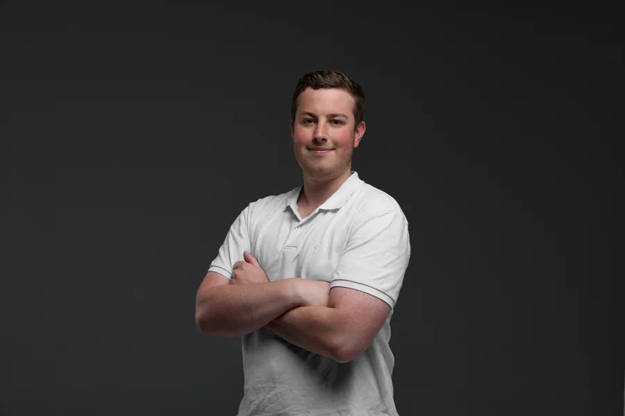 Studio headshot of Blake Harkness, arms crossed, in a white polo on a dark grey background