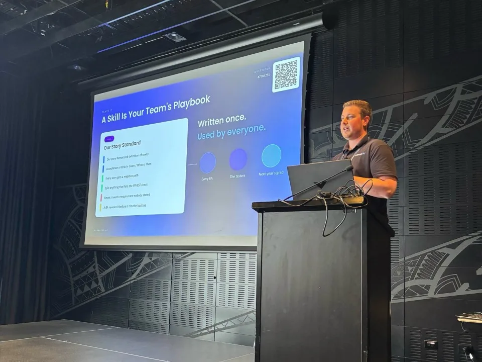 Blake Harkness at the lectern at the BA Big Day In 2026 at Tūranga in Christchurch, beside his slide A Skill Is Your Team's Playbook