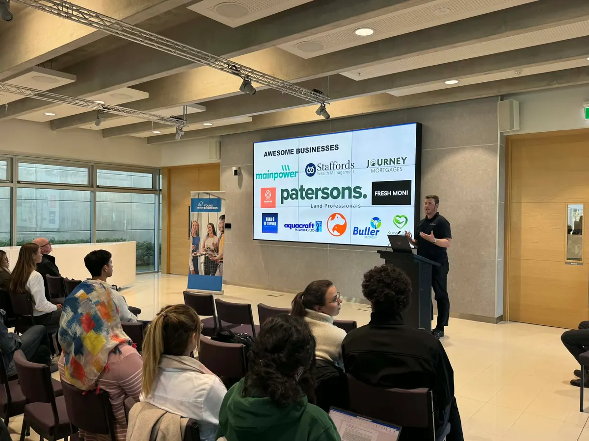 Blake Harkness at the lectern at the University of Canterbury in front of a slide of the businesses he works with, including Patersons, MainPower and Buller Electricity