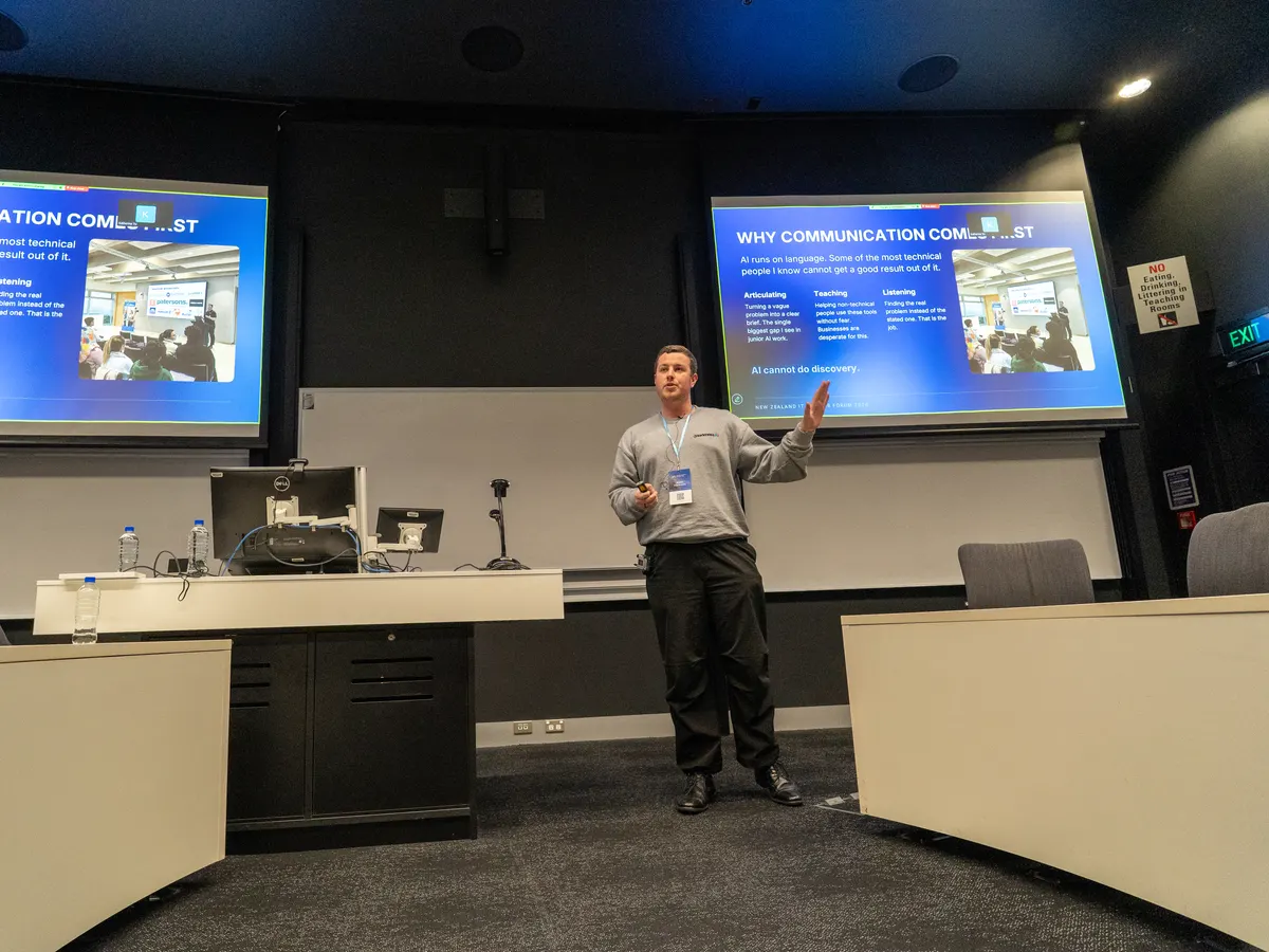 Blake Harkness on stage at the NZ IT & AI Job Forum at the University of Auckland, between two screens showing the slide Why Communication Comes First