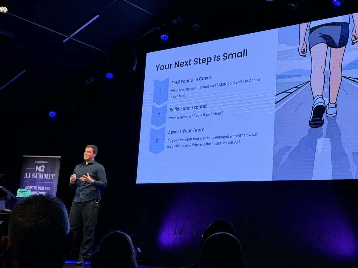 Blake Harkness on stage at the M2 AI Summit in Christchurch, beside the slide Your Next Step Is Small: find your use cases, refine and expand, assess your team